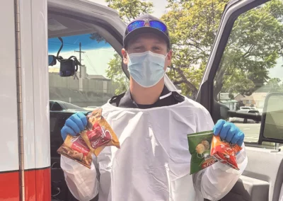 An EMS worker wearing PPE holds up snacks in front of an ambulance