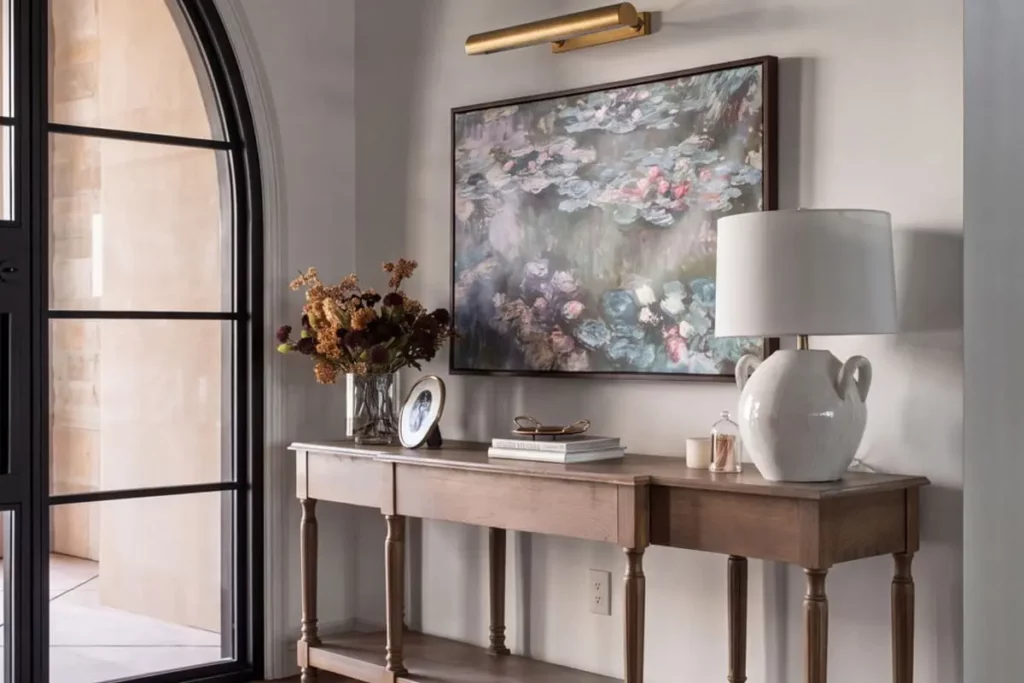 An entryway table with books, a vase of flowers, and a lamp with a large painting on the wall.